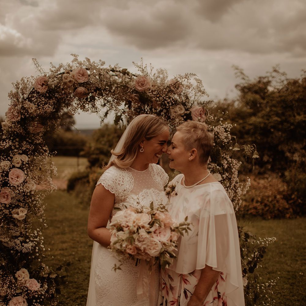 Bride in a lace wedding dress with her mum in front of a pink rose and white gypsophila flower arch 