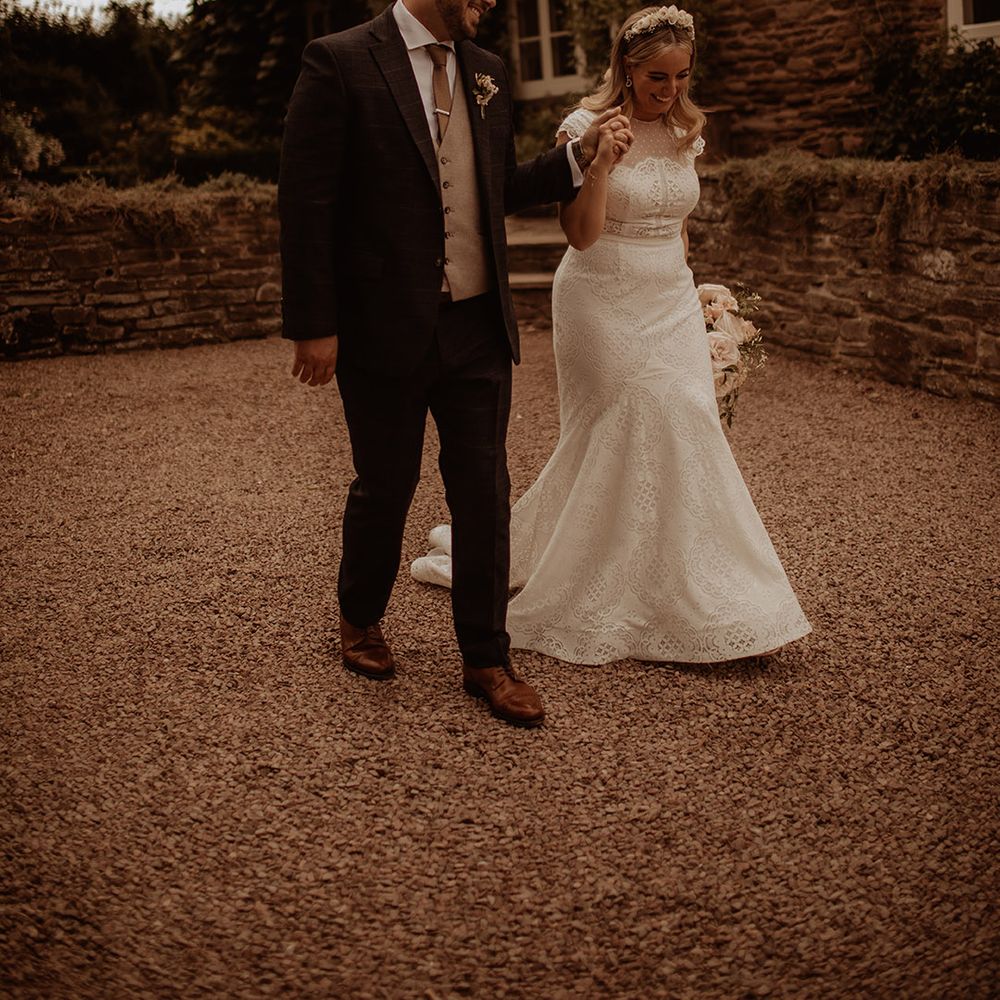 Groom in a three piece wedding suit holding hands with his bride in a lace fishtail wedding dress with cap sleeves