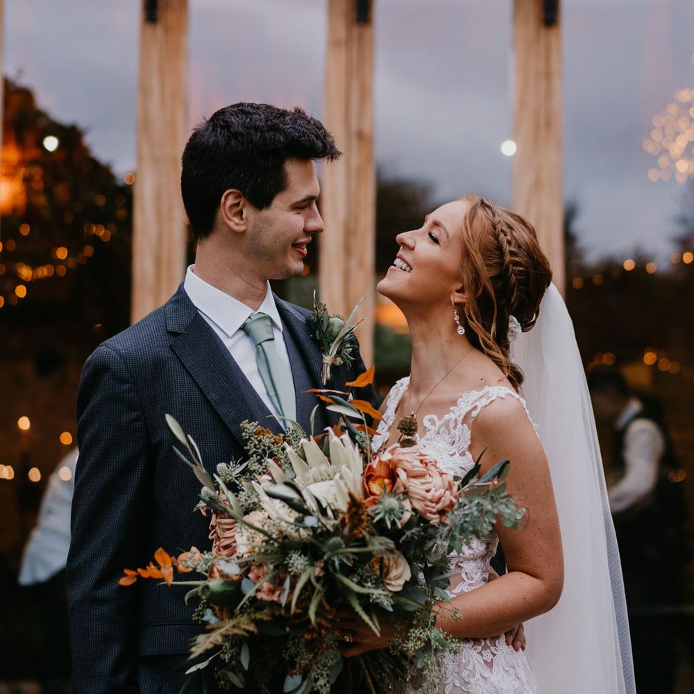 Bride with braided updo and veil holding large mixed bridal bouquet smiles at groom in dark suit and light grey tie outside barn