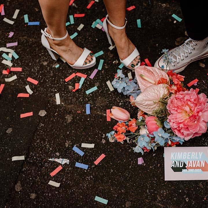 Bride in white platform shoes and groom in Converse high tops standing next to a pink and blue wedding bouquet 