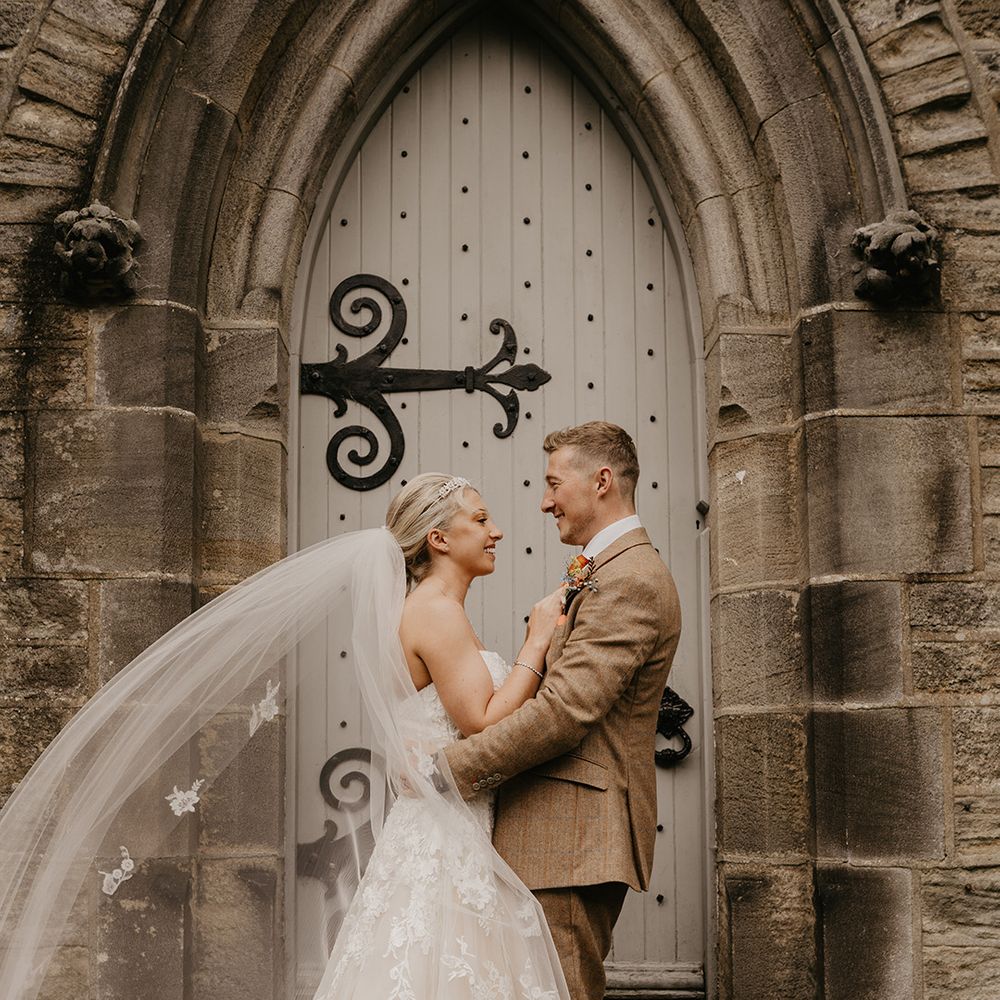blonde bride in strapless sweetheart lace wedding dress and embroidered veil with groom in beige suit in front of door