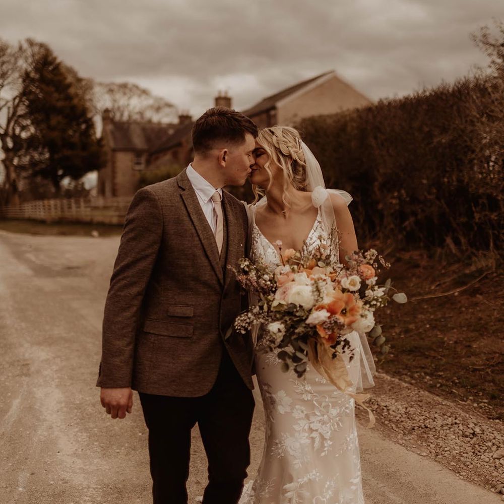 Groom in three piece brown suit kisses bride in lace Made With Love wedding dress as they walk around Eden Barn