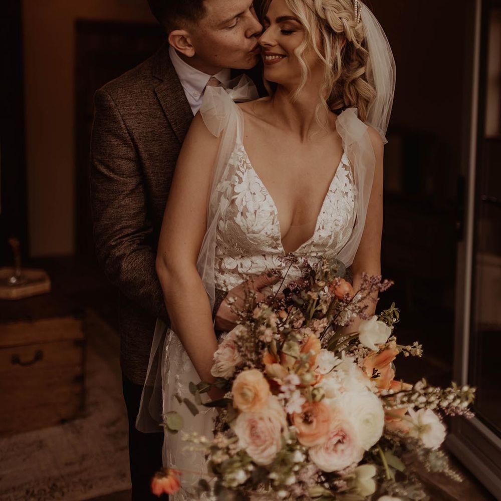 Groom in brown suit kisses the bride's cheek wearing Made With Love wedding dress and holding an autumnal wedding bouquet 