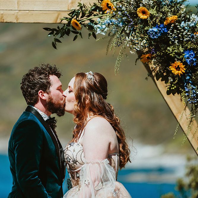 Bride & groom kiss beneath wooden arch finished with sunflower florals in front of the sea in Cornwall 
