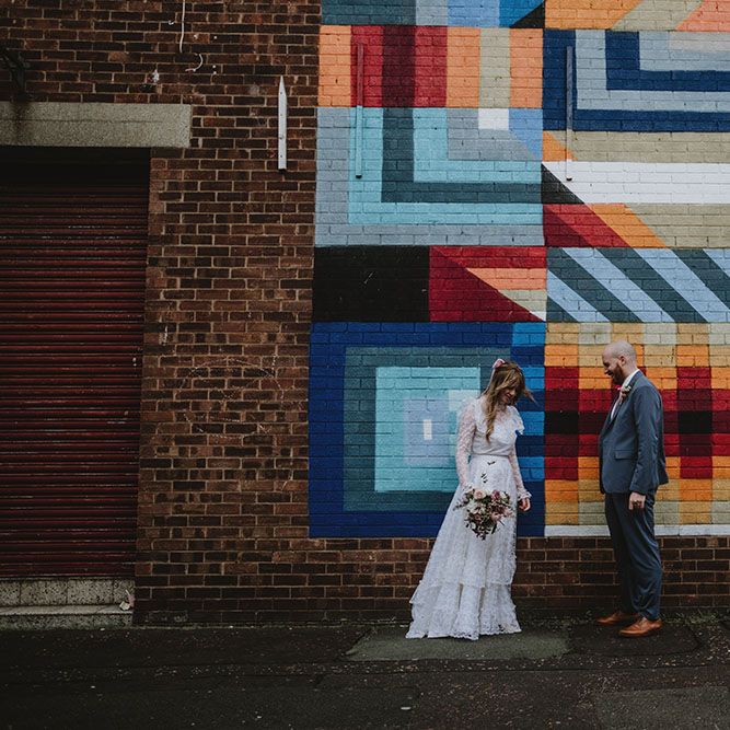 Bride wearing vintage lace wedding dress and pink bow hair accessory stands beside her groom in front of brightly coloured brick wall in Liverpool