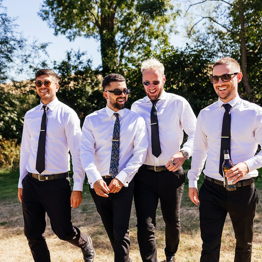 Groom walks with his groomsmen who wear white shirts and black ties with sunglasses outdoors 