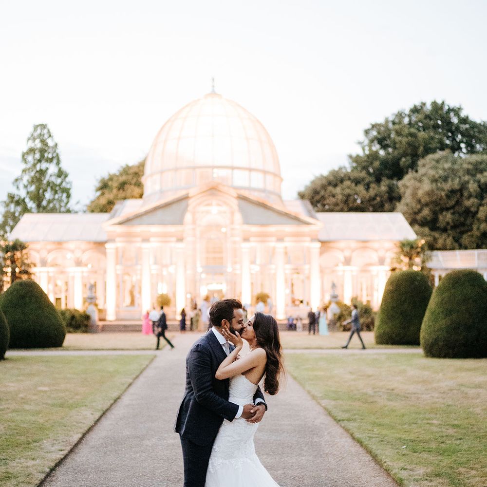 Bride & groom kiss outdoors in front of the brightly lit Great Conservatory at Syon Park