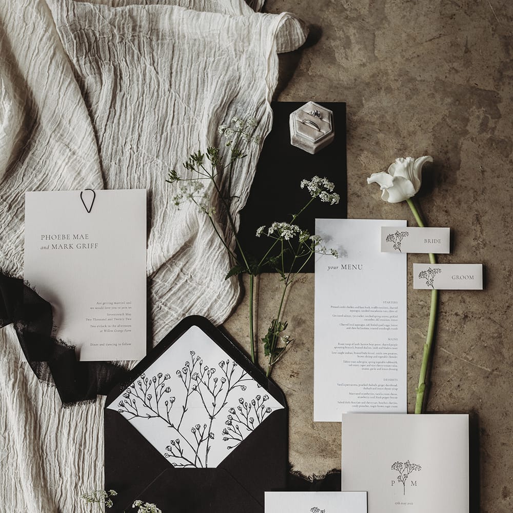 Wedding stationery suite flat lay with black and white invites, menus, RSVPs and envelope with white flowers and a ring box