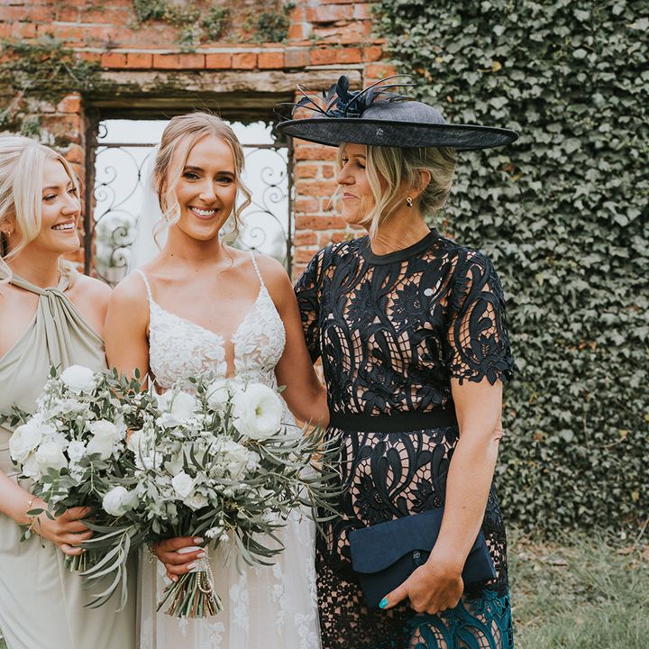 Bride in a floral lace wedding dress and bridesmaid in sage green dress with the mother of the bride in a black lace dress and hat
