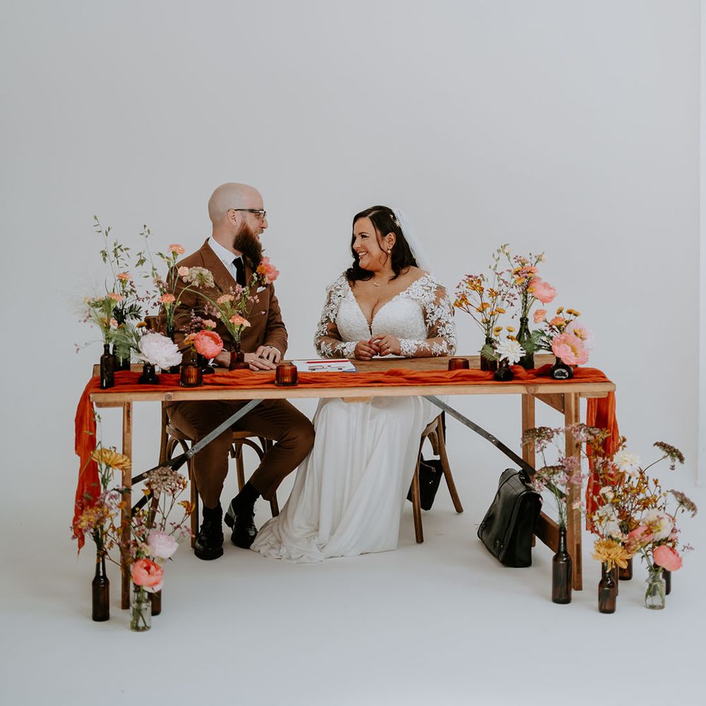 Bride & groom sit at wooden table complete with orange table runner surrounded by small floral arrangements 