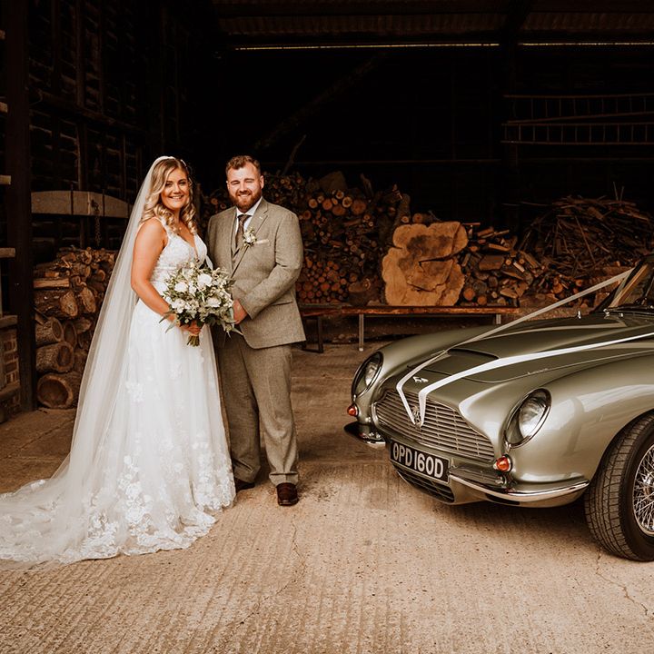 The bride and groom pose next to their Aston Martin grey wedding car transport 
