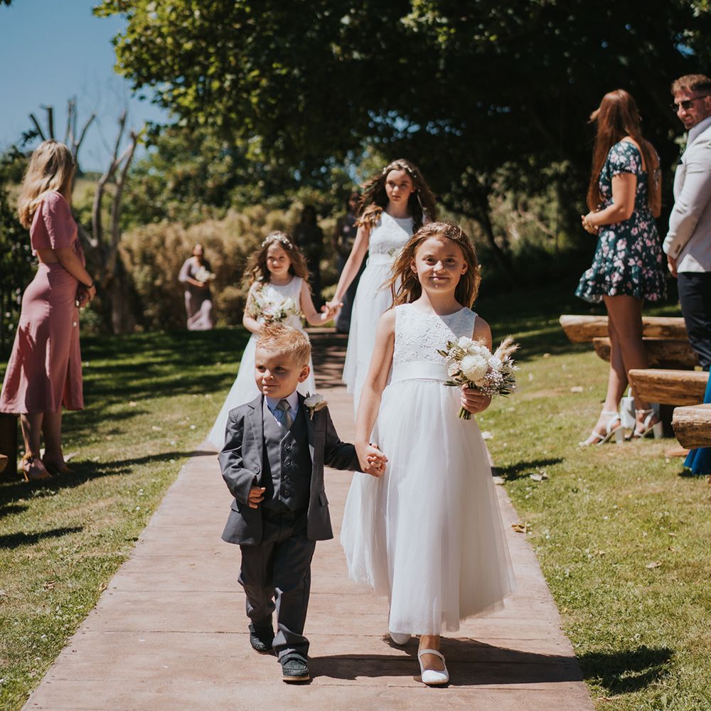 Flower girls in matching white dresses walk down the aisle with small floral bouquets and page boy in three piece suit