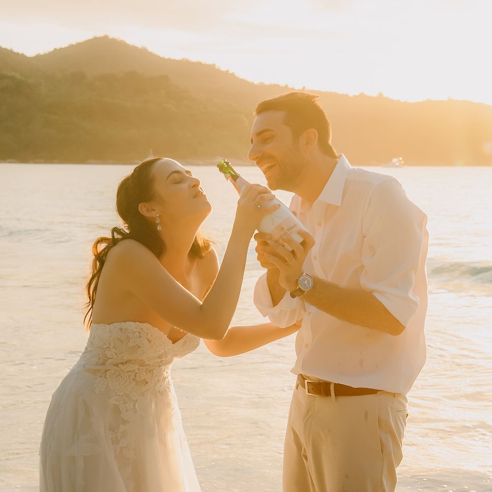 Bride in off the shoulder lace wedding dress with sweetheart neckline and lace floral details and groom in crisp white shirt, light coloured chinos and tan leather belt doing champagne pop on the beach 