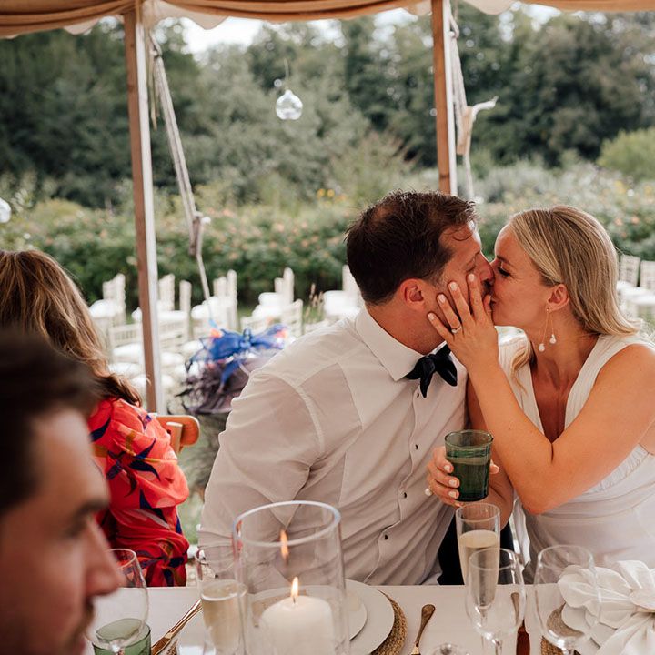 Groom in a white shirt sits with the bride as they share a kiss at their wedding reception in a marquee at Pythouse Kitchen Gardens 