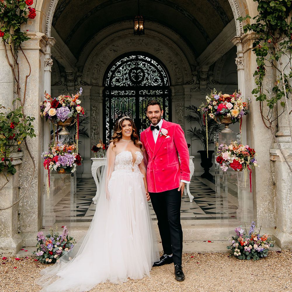 Bright and summery colourful wedding flowers decorate the altar space with the bride in a tulle wedding dress and groom in a bright pink jacket