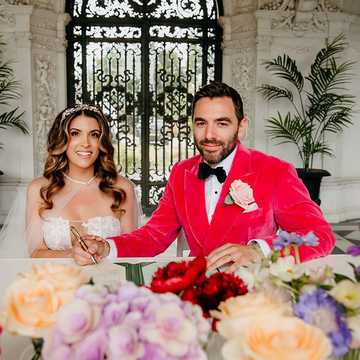 The bride in a strapless lace wedding dress sitting down with the groom in a bright pink jacket sign the register 