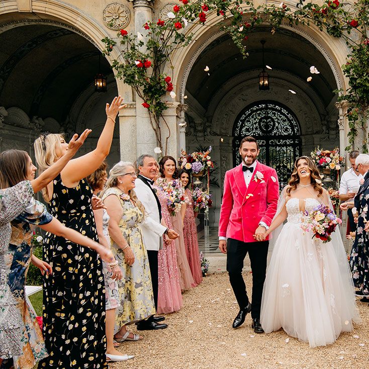 Confetti moment for bride in a sheer cape and groom in bright pink jacket as they leave their outdoor ceremony 