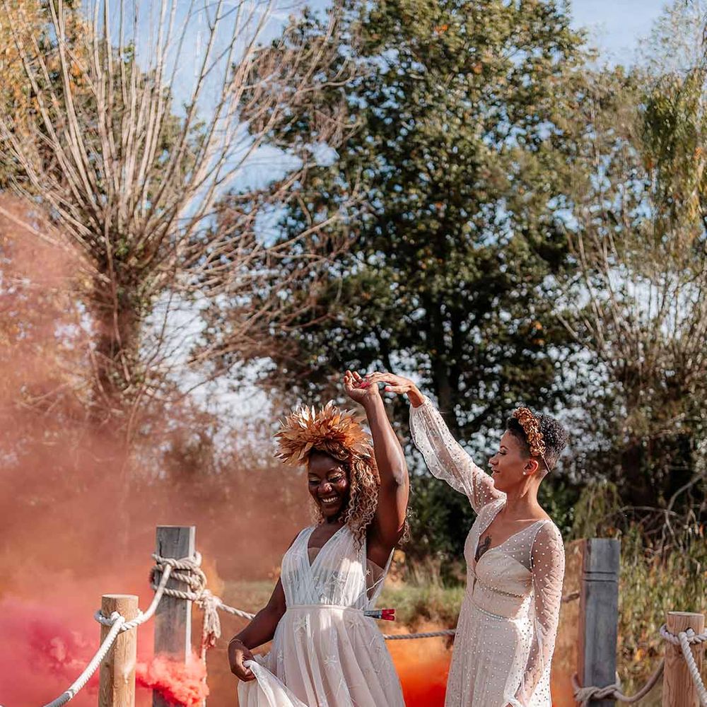 Brides in ethereal, boho wedding dresses and golden bridal crowns dancing in front of orange and pink smoke bomb on the dock of the river by Silchester Farm