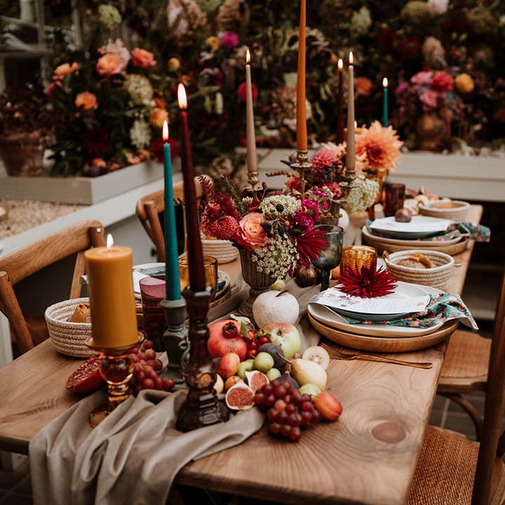 Neutral toned tablerunner on rustic wooden table, orange, green and red coloured glasses, earthenware, seasonal fruit and vegetables, red, brown and green tapered candles in gold candlesticks and clear vases with autumnal coloured flowers
