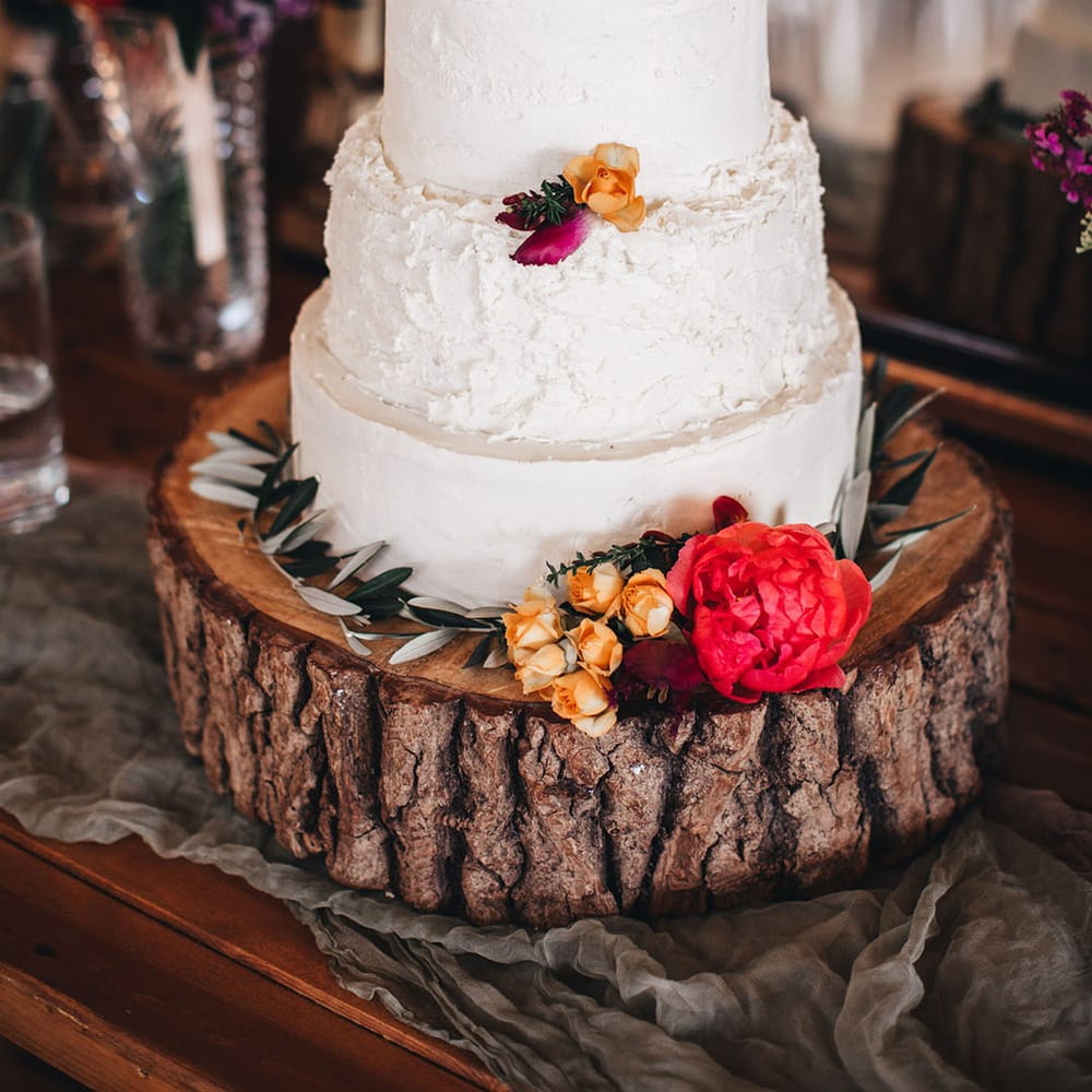 Three tier rustic wedding cake with white frosting on tree trunk cake stand with colourful flowers decorating the cake 