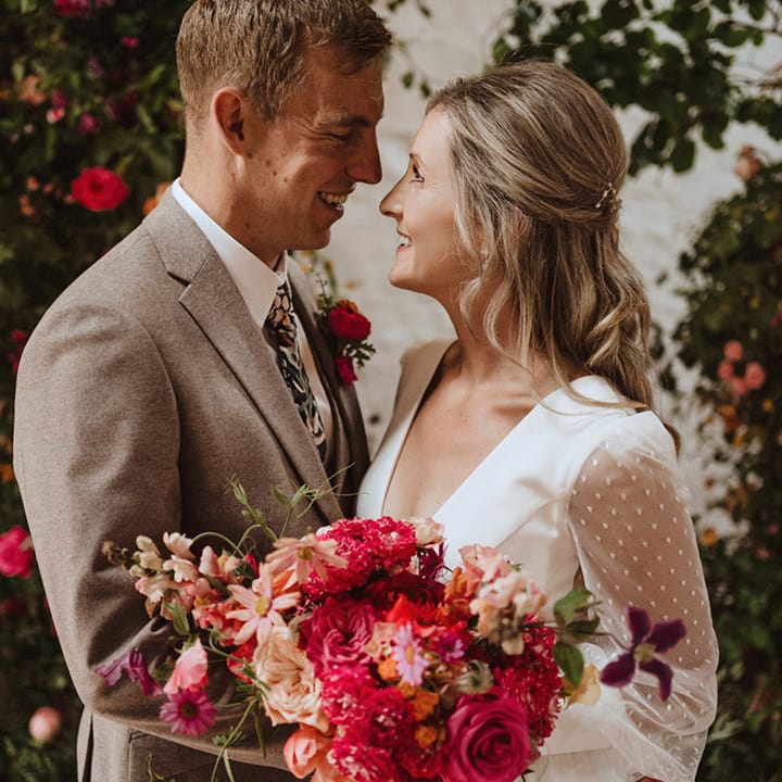 The groom in a neutral wedding suits gazes lovingly into the bride's eyes who wears a sheer long sleeve wedding dress with polka dots holding a pink peony and rose wedding bouquet