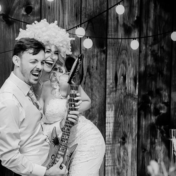 Groom posing with blow up guitar with the bride in a lace wedding dress wearing a fun wig for the wedding photo booth 
