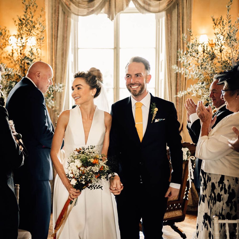 Groom in navy suit with yellow tie and tie clip walking back down the aisle with the bride in a plunging wedding dress carrying an orange bouquet 