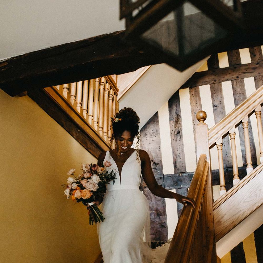 Bride in v neck sleeveless wedding dress with 3D floral applique train holding blush and orange garden roses, poppies and carnations, dried flowers and eucalyptus wedding bouquet walking down the stairs at Brinsop Court wedding venue