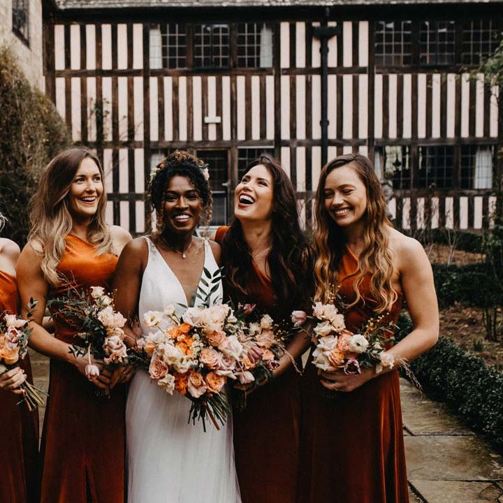 Bridesmaids in one shoulder burnt orange bridesmaid dresses holding blush and orange garden roses, poppies and carnations, dried flowers and eucalyptus wedding bouquets standing with bride in v neck sleeveless wedding dress with 3D floral applique train at Brinsop Court wedding