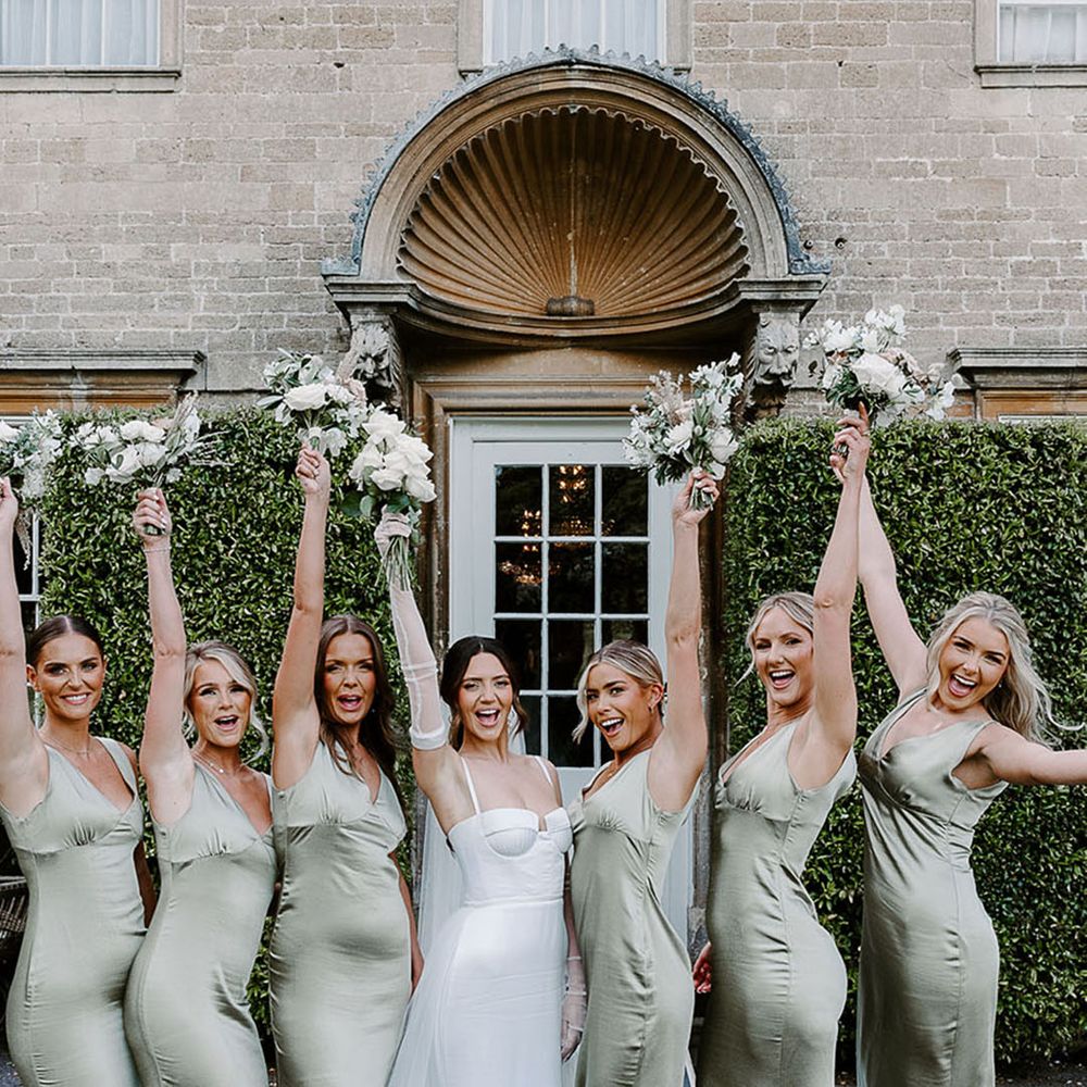 Bridal party with bridesmaids in sage green satin bridesmaid dresses with the bride holding white flower bouquets 