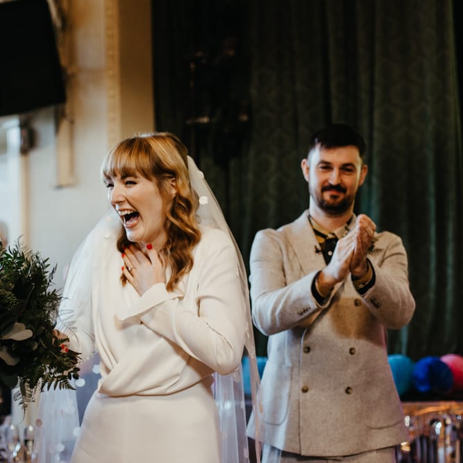 Smiley bride in a jumpsuit and appliqué veil holding a green foliage wedding bouquet with red nail polish