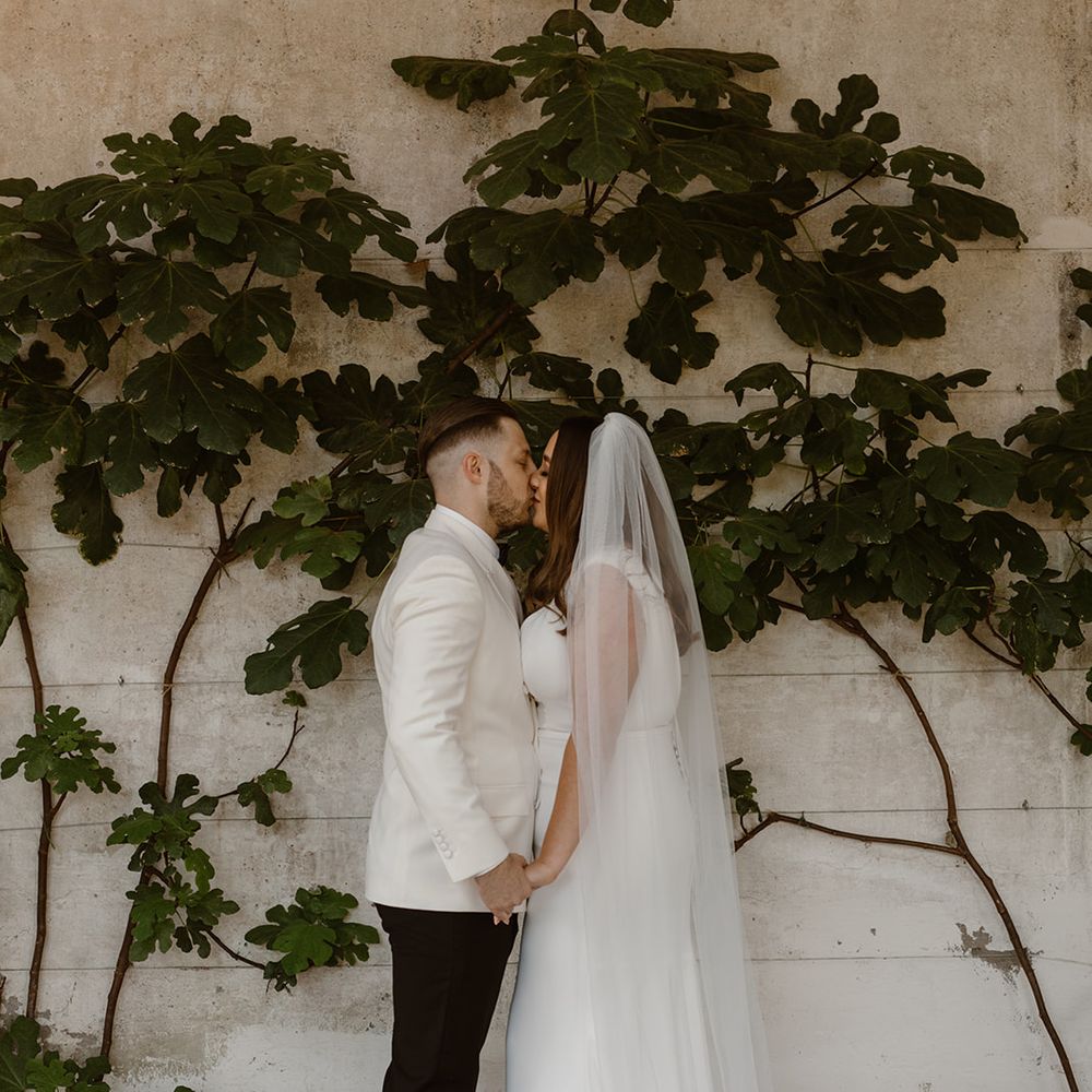 Bride in V neck Made With Love wedding dress and chapel length veil stands holding hands with groom in white double breasted tuxedo jacket