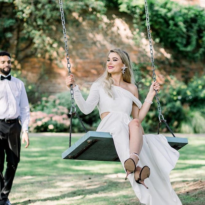 Stylish bride in a one shoulder wedding dress with long sleeve and black sandals sitting on a swing at Garthmyl Hall 