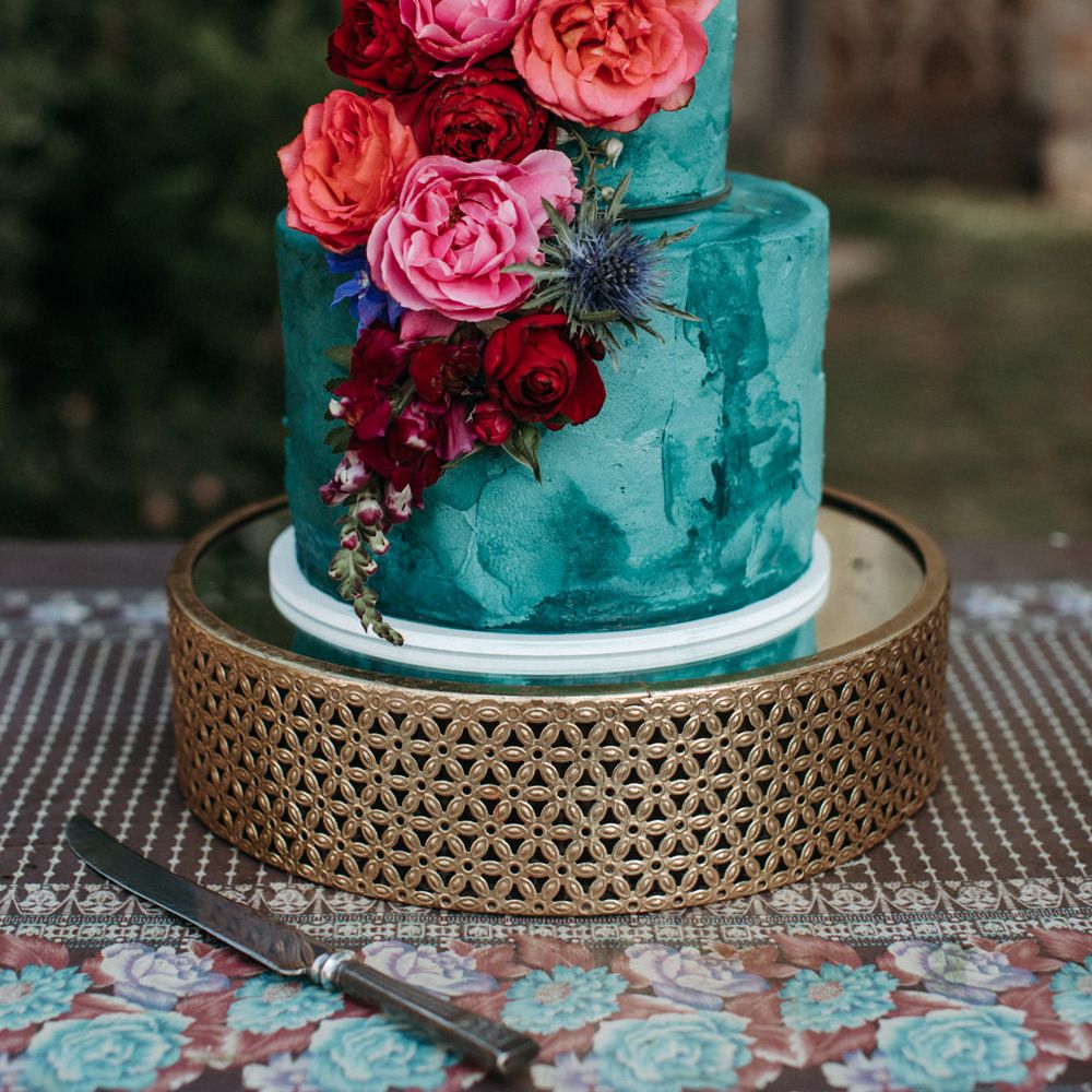 Two tier turquoise wedding cake on a gold cake stand with pink and red peony and rose cake decor