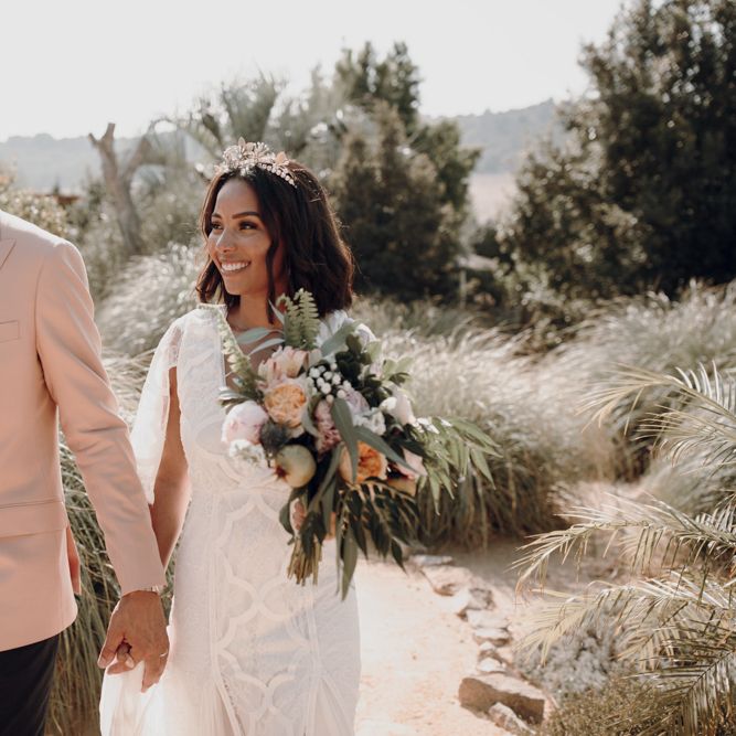 Groom in a pink double breasted jacket and floral bow tie holding hands with his bride in a lace boho wedding dress