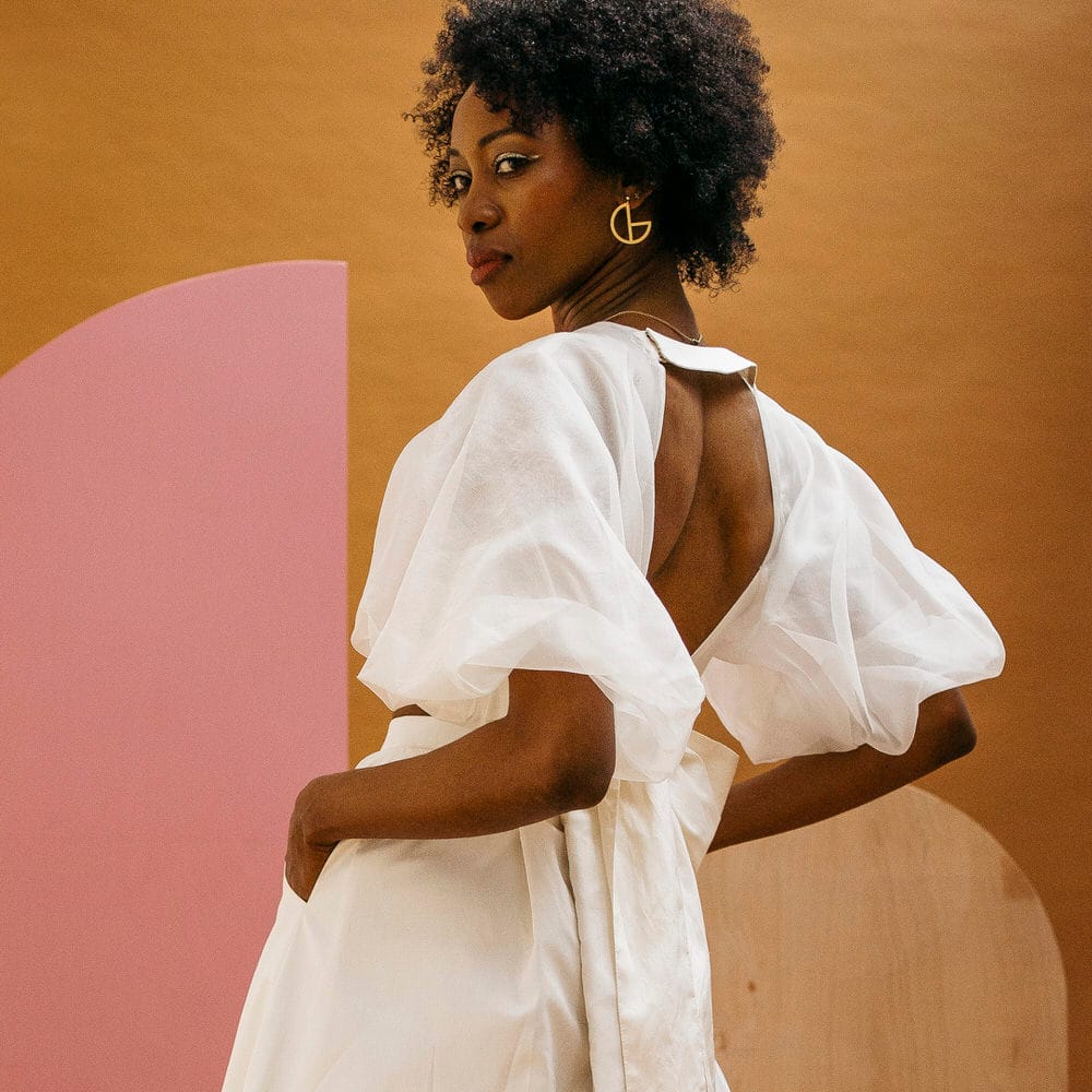 Bride with afro hair wearing puff sleeve, open back wedding separates with bow detail in front of a mustard colour backdrop