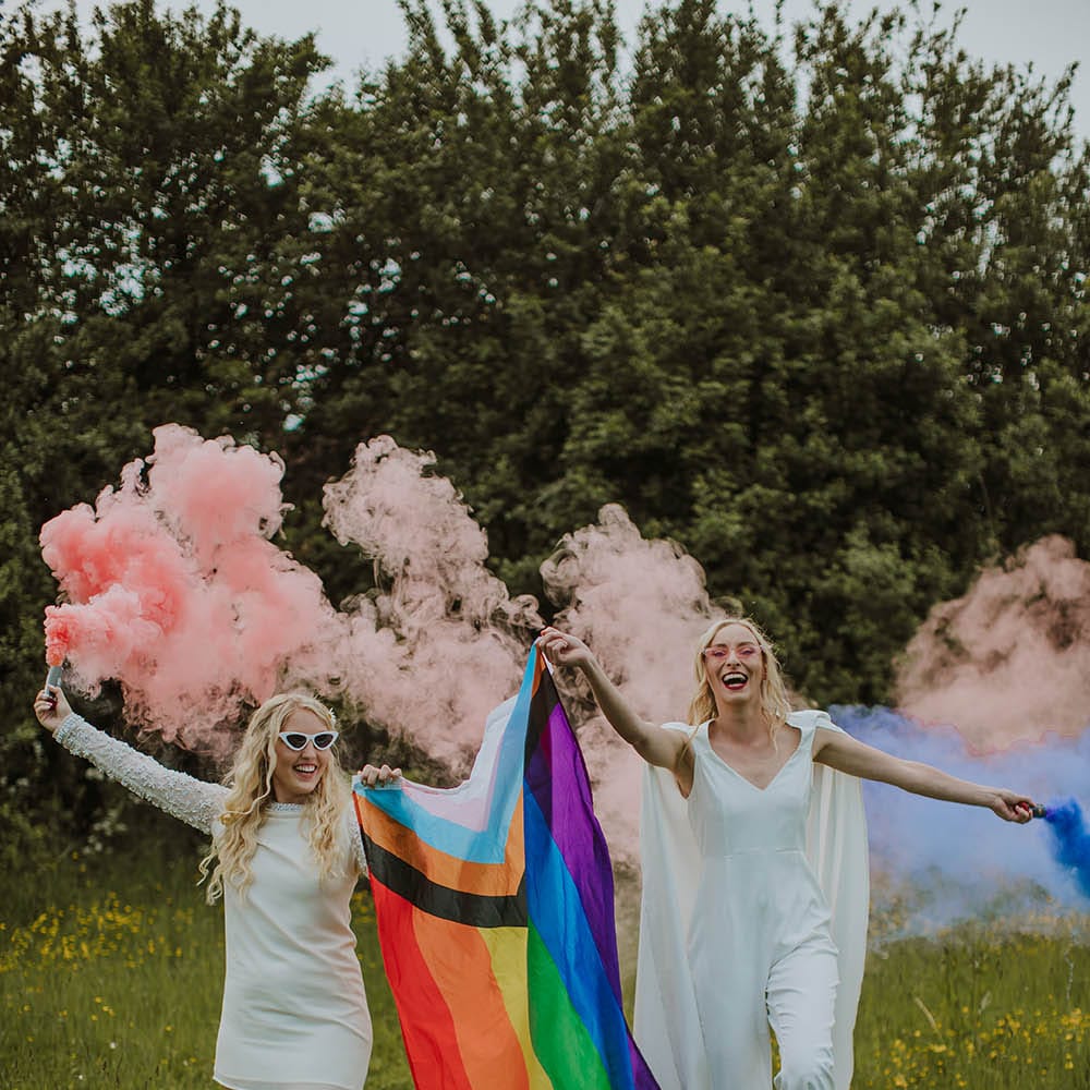 Two brides in a short wedding dress with long sleeves and bride in a jumpsuit and cape holding smoke bombs and a pride flag 