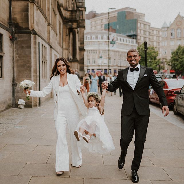 Family wedding portrait with bride and groom in black and white suits swinging their flower girl daughter in the air 