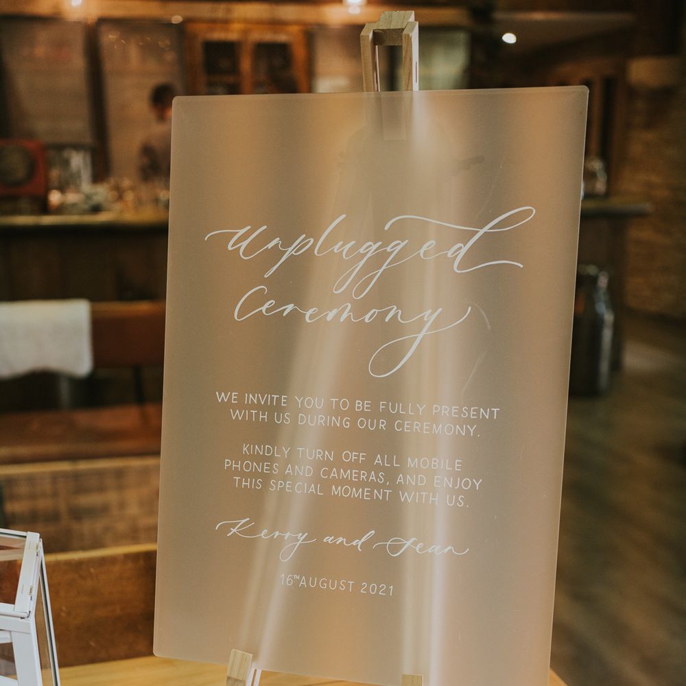 Frosted acrylic wedding sign reading 'unplugged ceremony' on wooden table with white flowers and green leaf decor for Tythe Barn wedding with barn wedding flowers