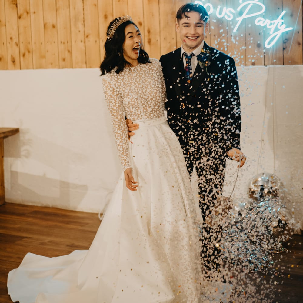 Bride in long sleeved applique wedding dress stands with groom in dark suit and floral tie as disco ball piñata confetti falls at rustic wedding with neon sign