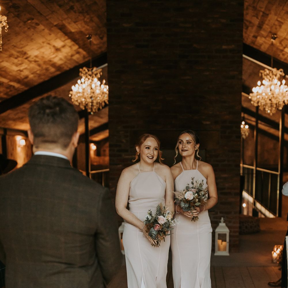 Two bridesmaids in light pink halterneck bridesmaid dresses holding mixed rustic bridal bouquets walk down barn aisle 