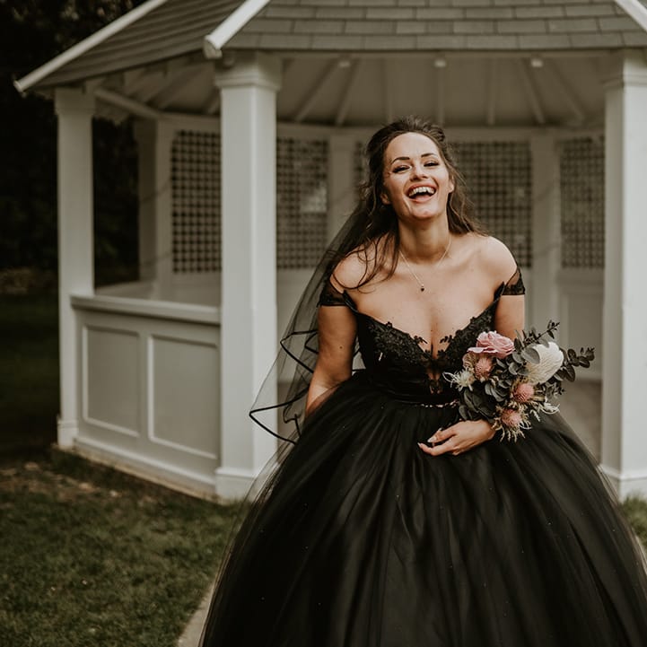 Bride laughs and wears black wedding gown with off the shoulder straps and holds floral bouquet 
