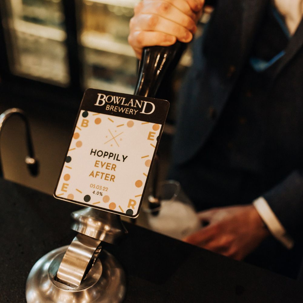 Groom in navy suit pulls a draft ale from the pump labelled 'Hoppily Ever After' during wedding reception at Holmes Mill