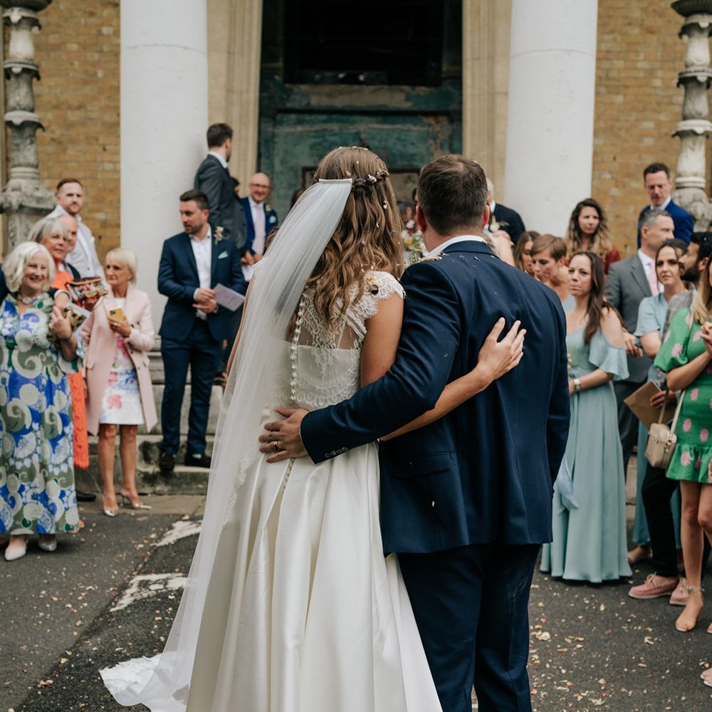 Bride with waterfall braid hairstyle and buttoned lace wedding top and pleated skirt standing with groom in navy blue 