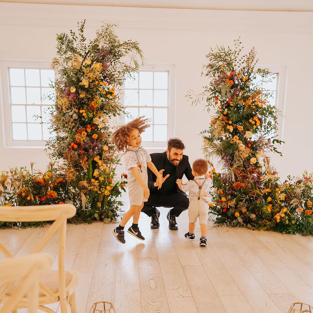 Groom in all black playing with page boys in floral shirts, braces and shorts in between orange and green column wedding ceremony flower arrangements 