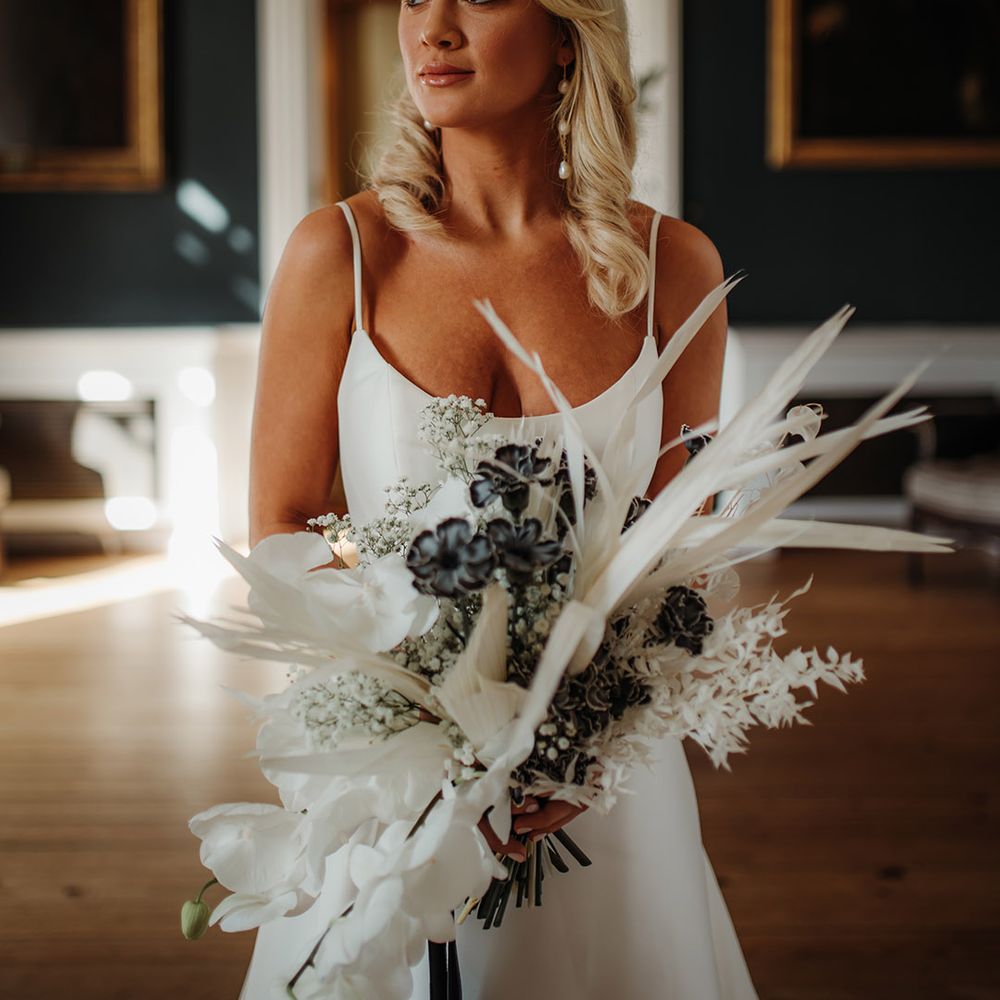 Blonde bride with long wavy hear wearing pearl earrings and holding a dried and fresh flower boqueut with orchids 