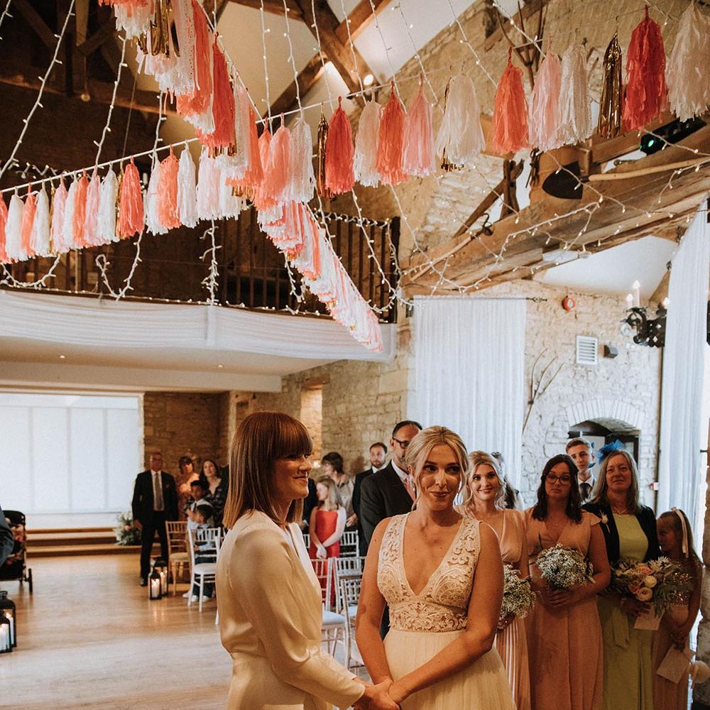 LGBTQI+ wedding ceremony at The Great The Barn with two brides holding hands at the altar as they exchange vows under a canopy of tissue bunting 