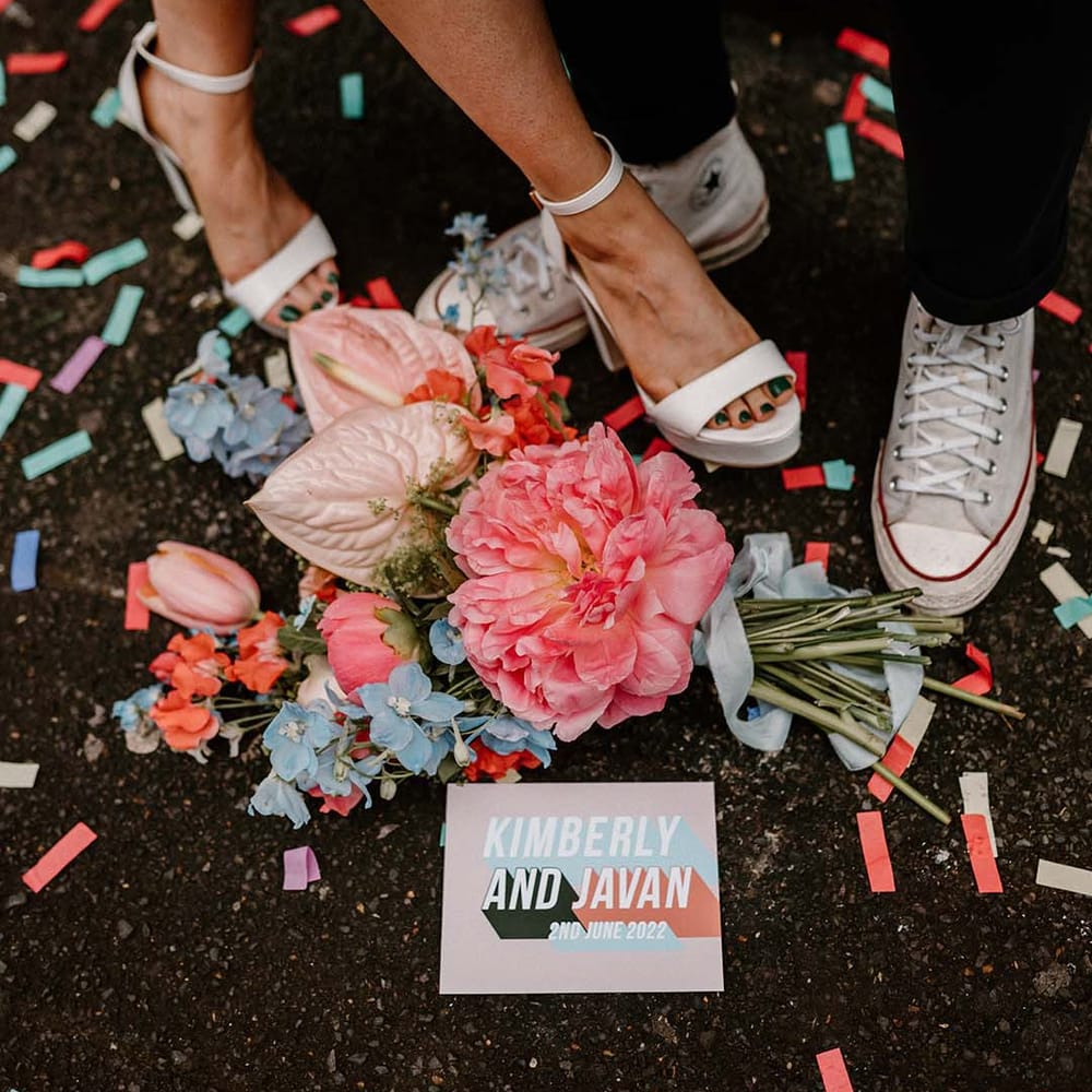 Coral pink peony, baby pink anthuriums and blue delphinium wedding bouquet resting on a floor sprinkled with confetti 