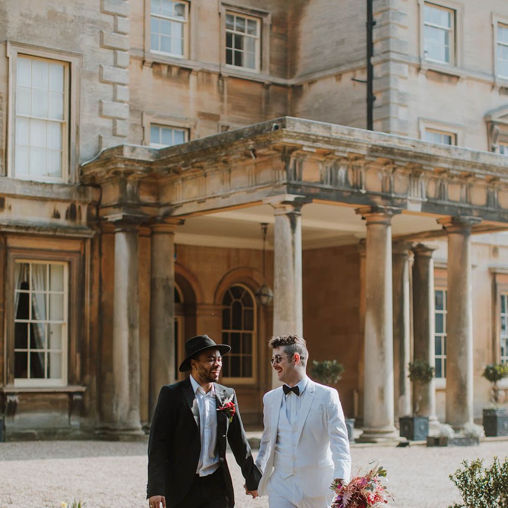 Groom in a two piece black suit, fedora hat and boots holding hands with his partner in a white three piece groom suit and white boots at Prestwold Hall Barns