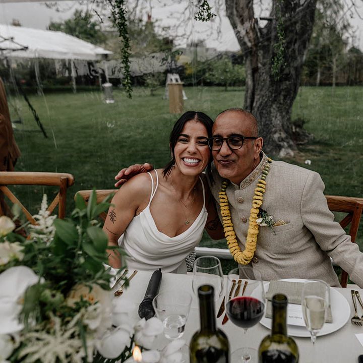 Indian bride in a slip wedding dress embracing her father at the top table of her clear glass wedding reception 